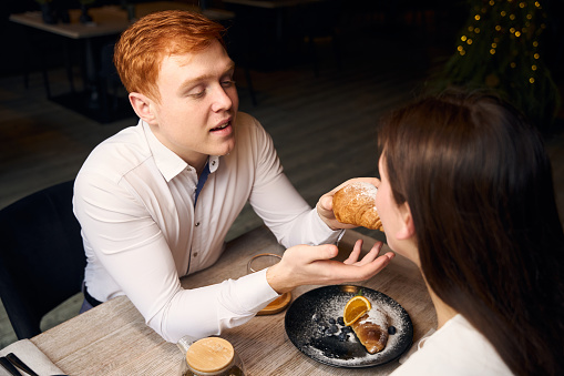 Caring red-haired guy treats his girlfriend with a croissant, the couple is located in a cozy cafe by the window