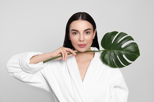 Woman in bathrobe holding leaf of monstera on light grey background. Spa treatment