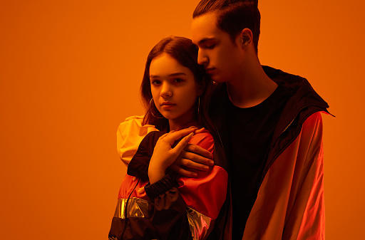 Serious teen man hugging female model wearing stylish jacket in studio against orange background