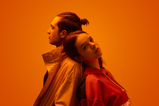 Side view of young couple of teen boyfriend and girlfriend in trendy clothes, standing back to back on orange background in studio with neon light