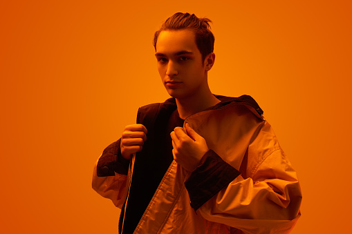 Young male looking at camera and adjusting stylish oversize jacket while standing under bright neon illumination against orange background