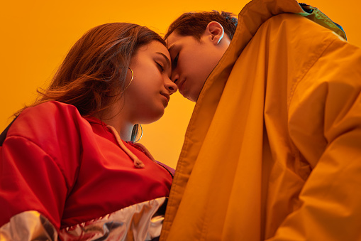 From below male teenager in stylish coat kissing girlfriend against yellow background under neon illumination