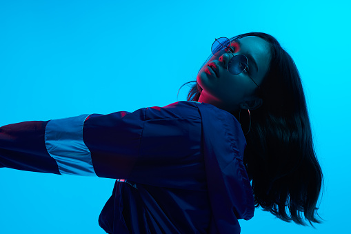Side view of cool emotional young female hipster in glasses looking at camera in studio with neon light on blue background