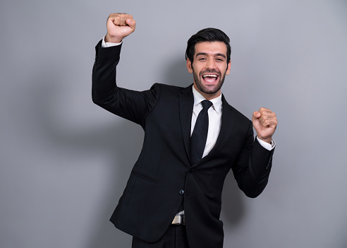 Excited and happy businessman dressed in black formal suit, raise his arm in celebratory gesture on isolated background, after successful job promotion, energy and joy as business winner. Fervent