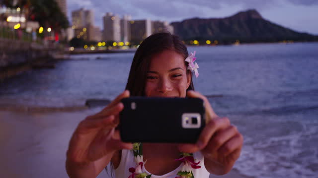 Happy young woman photographing using smartphone at Waikiki Beach. Smiling female tourist is enjoying vacation at island in Honolulu wearing orchid lei garland taking photo.