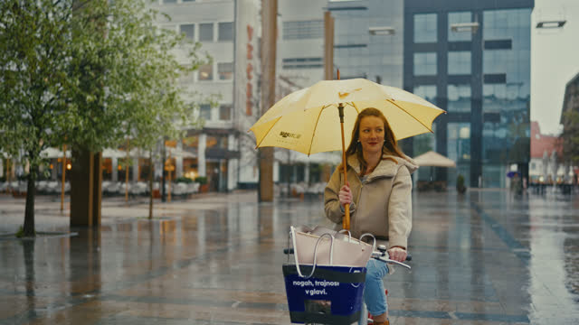 SLO MO Young woman with a yellow umbrella cycling in the rain in the city