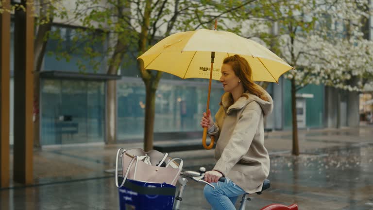 SLO MO Young woman with a yellow umbrella cycling in the rain in the city