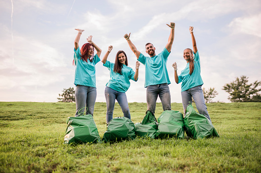 Spontaneous image of a small group of active and healthy living, people, young adults, taking care, voluntarily, of nature, cleaning park area, with a big green garbage bags. They're wearing a protective gloves and all uniformed blue t-shirts. Having fun while standing in front of bags, feeling good for doing the right thing, smiling at the camera