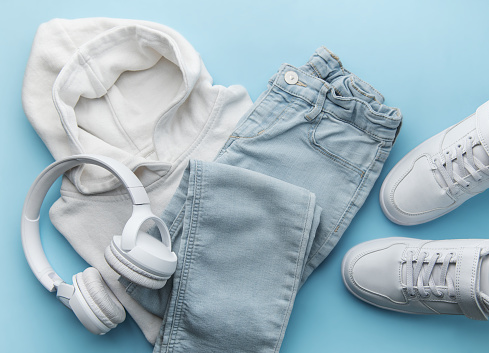 Childs clothing, accessories, footwear,  sweatshirt,  jeans,  sneakers, headphones on blue background. Outfit for teens. Top view, flat lay. Trendy colors