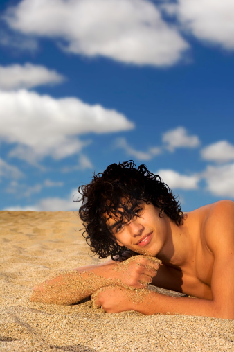 Male teenager with dark curly hair and beautiful face lying on the sand at beach