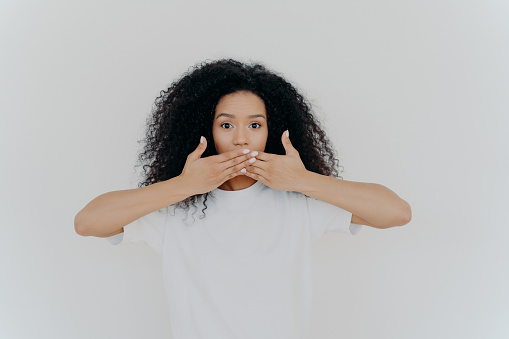 Horizontal shot of surpised dark skinned woman covers mouth with both palms, tries not to speak, dressed in casual mockup t shirt, poses against white background. People, secretness concept.