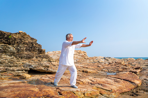 Asian senior man practicing meditation with nature on rocky coastal hill. Retirement elderly people do outdoor relaxing tai chi yoga exercise at summer sunset. Mental health care motivation concept.
