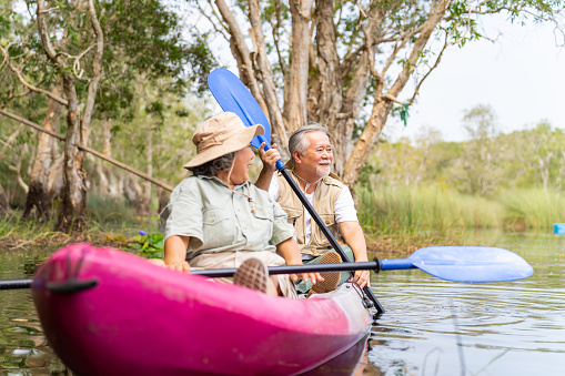 Happy Asian senior couple kayaking together in the lake at mangrove forest on summer vacation. Retired elderly people man and woman have fun outdoor lifestyle travel nature and rowing a boat in the river.