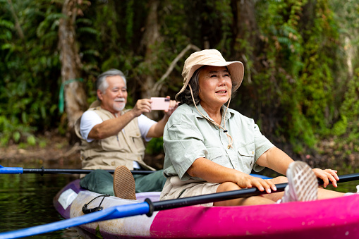 Happy Asian senior couple using mobile phone taking picture together during kayaking in the river at mangrove forest. Retired elderly man and woman enjoy outdoor lifestyle travel nature on summer vacation.