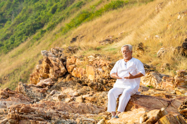 Asian senior man practicing meditation with ocean nature on rocky coastal hill at summer sunset. Retirement elderly people do outdoor relaxing yoga exercise. Mental health care and motivation concept.
