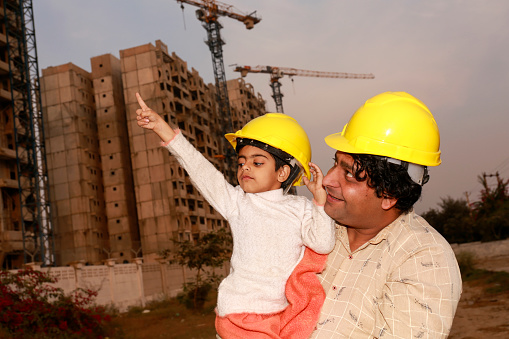 Cheerful father and elementary age daughter of Indian ethnicity standing portrait near construction site wearing protective helmet.