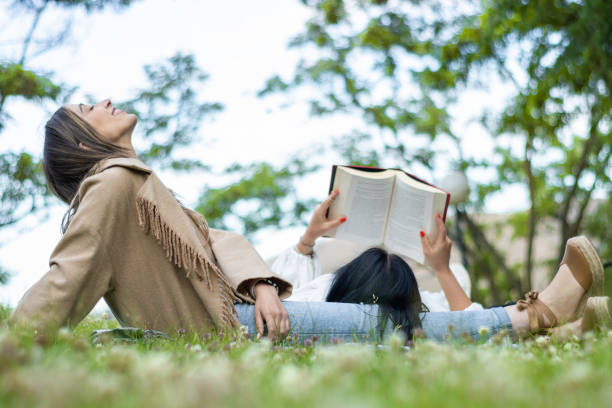 Side view of two relaxed female friends enjoying a day together in the park while getting some fresh air and reading a book.