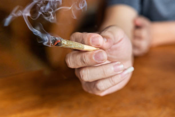 istock Finger holding lighted cannabis cigarette with smoke in dark room, with selective focus on cannibis ash and fire. 1482371884
