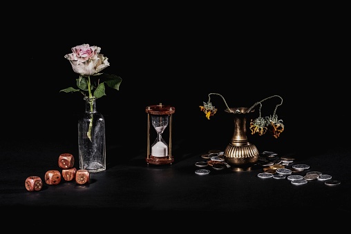 A still life shot of a timeglass, wilting flowers, money, and dice on a black background