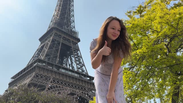 a beautiful teenage girl looks into the frame leaning against the backdrop of the Eiffel Tower She smiles and seems to be showing come here great advertisement for a trip to Paris