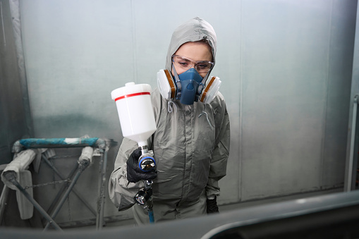 Caucasian woman auto technician wearing protective workwear painting car body element during repair work in paint chamber