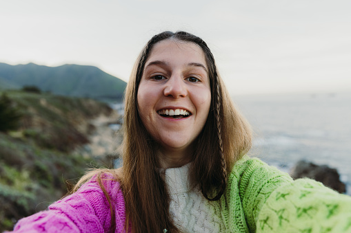 Self-portrait of female with long hair wearing colourful knitted sweater exploring the scenic mountain coast and making selfie on Monterey peninsula in the USA during sunny spring day