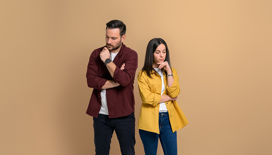 Disappointed young boyfriend and girlfriend dressed in casuals with hands on chins looking down. Unhappy couple contemplating after an argument while standing over beige background