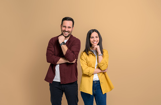 Portrait of cheerful attractive boyfriend and girlfriend dressed in casuals looking at camera. Smiling young couple touching chins and posing happily on isolated beige background