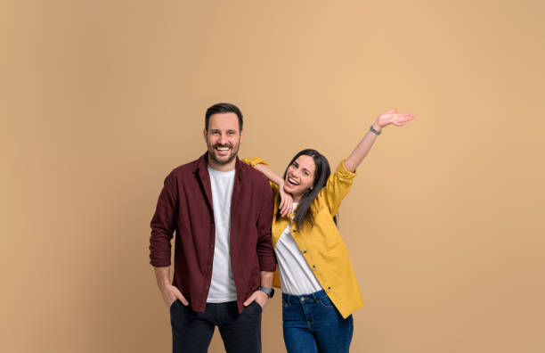 alegre joven novia con la mano levantada gritando y apoyándose en el novio guapo. retrato de una atractiva pareja vestida con camisas posando alegremente sobre fondo beige - affectionate adult beautiful attractive female fotografías e imágenes de stock