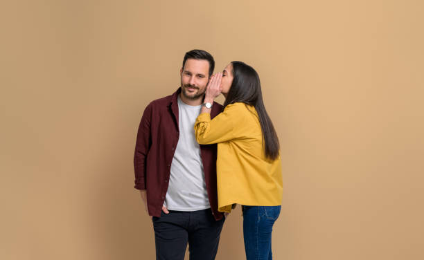 istock Young girlfriend whispering her desires into handsome boyfriend's ear while standing isolated over beige background. Attractive couple dressed in casuals gossiping together 1482421470