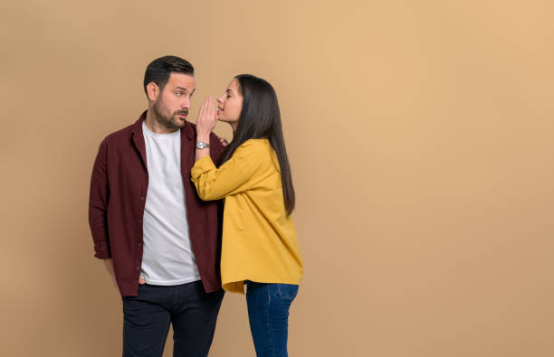 istock Serious boyfriend looking at girlfriend whispering secret into his ear while standing isolated over beige background. Young man and woman telling gossips to each other 1482421484