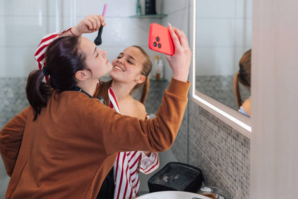 Two teenage best friends are bonding over their shared love of makeup in the bathroom. They chat and laugh as they apply their favorite products, and offer each other tips and tricks to enhance their looks