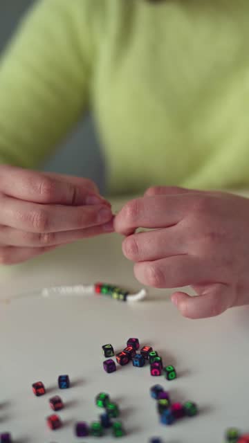 A little Girl with long hair and a yellow sweater Weaves Beaded jewelry. Close-up. Vertical video