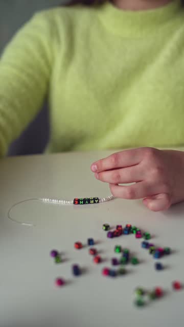 A little Girl with long hair and a yellow sweater Weaves Beaded jewelry. Close-up. Vertical video