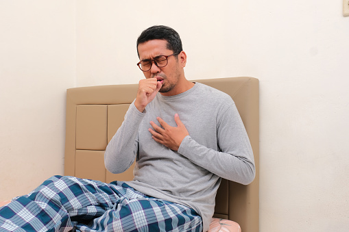 Adult Asian man sitting on his bed suffering bad cough
