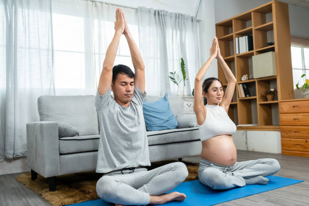 Asian men and women are Husband and wife with pregnant wives wearing open belly shirts. sitting in a yoga pose with hands raised above their heads to relax at leisure in middle of the living room. los hombres y mujeres asiáticos son marido y mujer con esposas embarazadas que usan camisas de vientre abierto. sentarse en una postura de yoga con las manos levantadas por encima de sus cabezas para relajarse en el medio de la sala de estar.