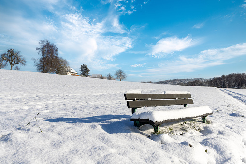 A lone wooden bench covered with a thick layer of soft white snow stands on a hillside against a frozen dense dark forest, against a blue sky on a clear sunny winter day