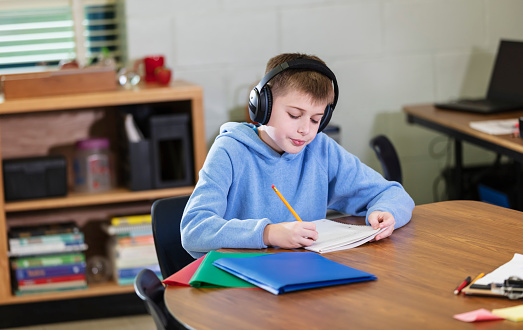 An elementary student sitting at a table in school, studying, writing in a notebook. The 11 year old boy is wearing noise-cancelling headphones to block out distracting sounds. Headphones may help students with ADHD or other learning disabilities to focus.
