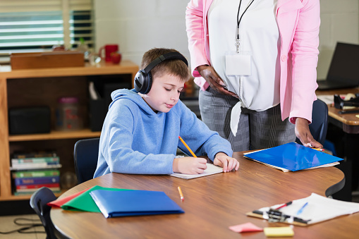 An elementary student sitting at a table in school, writing in a notebook. The 11 year old boy is wearing noise-cancelling headphones to block out distracting sounds. Headphones may help students with ADHD or other learning disabilities to focus. His teacher, a pregnant, African-American woman, is standing next to him, only her midsection visible.