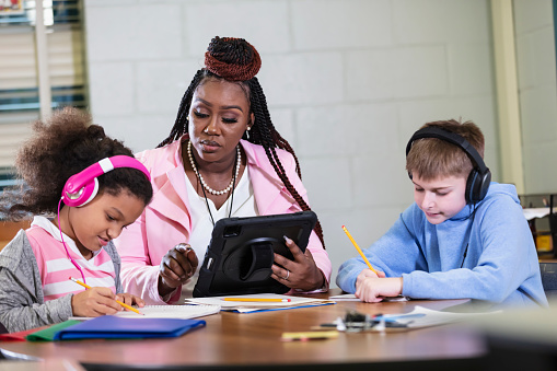 A teacher helping two elementary students sitting at a table in school, writing. The students are wearing noise-cancelling headphones to block out distracting sounds. Headphones may help students with ADHD or other learning disabilities to focus. The teacher, a pregnant, African-American woman, is sitting between the children, looking toward the 9 year old multiracial girl.