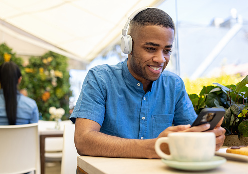 Happy African American man using his cell phone at a cafe while listening to music with headphones
