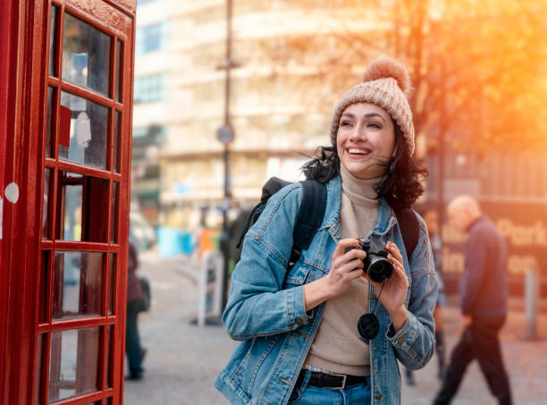 outdoor-porträt einer frau, die kamera gegen rote telefonzelle in einer englischen stadt verwendet - pay phone telephone people women stock-fotos und bilder