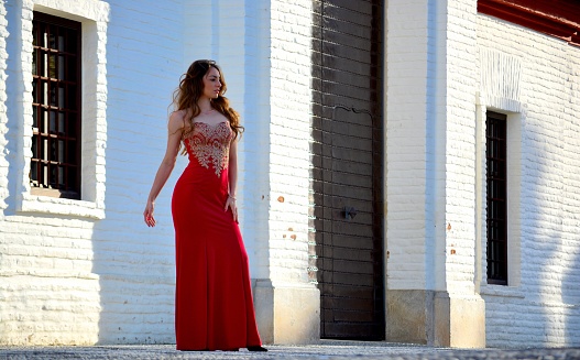 Elegant and standing young brunette woman dressed in a long oriental red dress in front of a typical white stone building in Spain, Europe