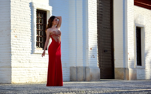 Elegant and standing young brunette woman dressed in a long oriental red dress in front of a typical white stone building in Spain, Europe