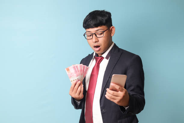 Portrait of young Asian business man in casual suit looking at smartphone surprised while holding thousand rupiahs. Isolated image on blue background stock photo