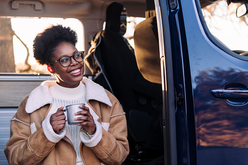 young black woman laughing happy at sunset sitting in a camper van with a cup of coffee in her hand, concept of van life and weekend getaway, copy space for text