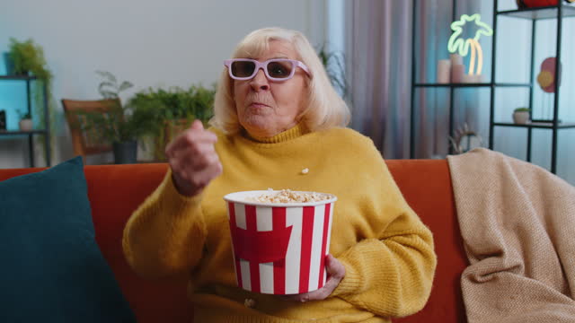 Grandmother woman on sofa eating popcorn, watching interesting TV serial, sport game online at home