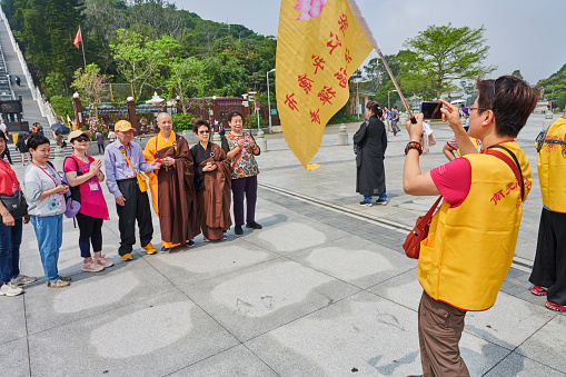 Ngong Ping, Hong Kong - April 15, 2023: a group of chinese tourists taking a photo with a buddhist monk at the Po Lin Monastery.