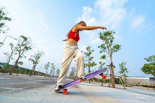 Cool Asian girl skating on longboard skate at public park on summer holiday vacation. Stylish young woman enjoy and fun urban active lifestyle practicing outdoor sport skateboarding on city street at sunset.