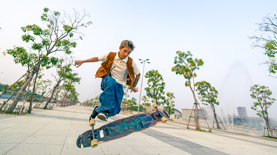 Cool Asian man skating on longboard skate at park on summer holiday vacation. Stylish young guy enjoy and fun urban active lifestyle practicing outdoor sport skateboarding on city street at sunset.